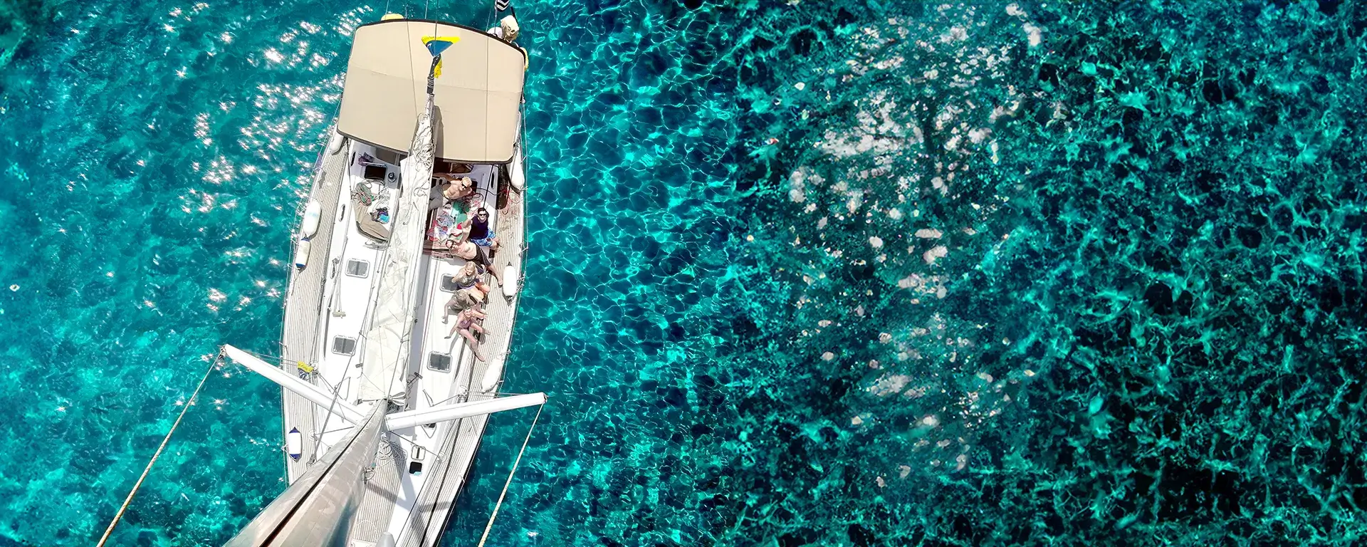 Aerial view of a luxurious sailboat floating on crystal-clear turquoise waters, with a group of people relaxing on deck. The yacht's white deck contrasts beautifully with the shimmering sea, creating a perfect summer vacation scene.