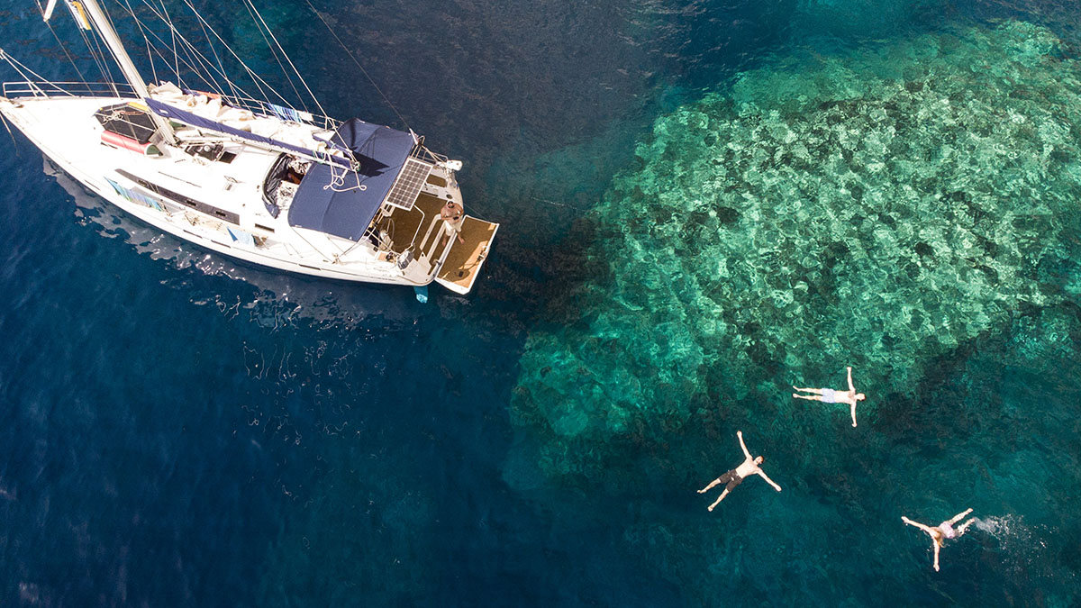 Aerial view of three people swimming and floating leisurely near a white sailboat anchored in crystal-clear turquoise waters, creating an idyllic scene for relaxation and vacation