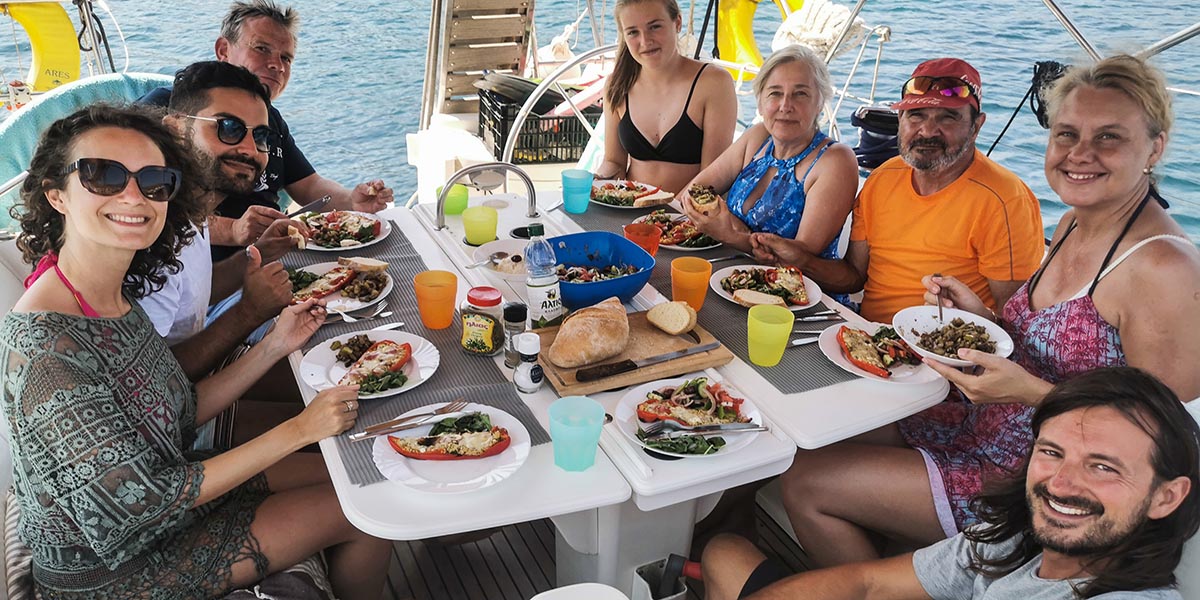 Group of happy people enjoying a meal together aboard a sailboat, smiling at the camera with fresh food, colorful plates, and calm blue waters in the background, creating a joyful sailing vacation atmosphere