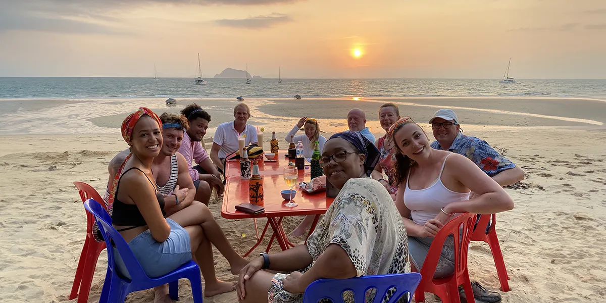 Group of friends happily gathered around a beachside table, smiling and enjoying drinks together as the sun sets over the ocean, creating a warm and inviting tropical atmosphere