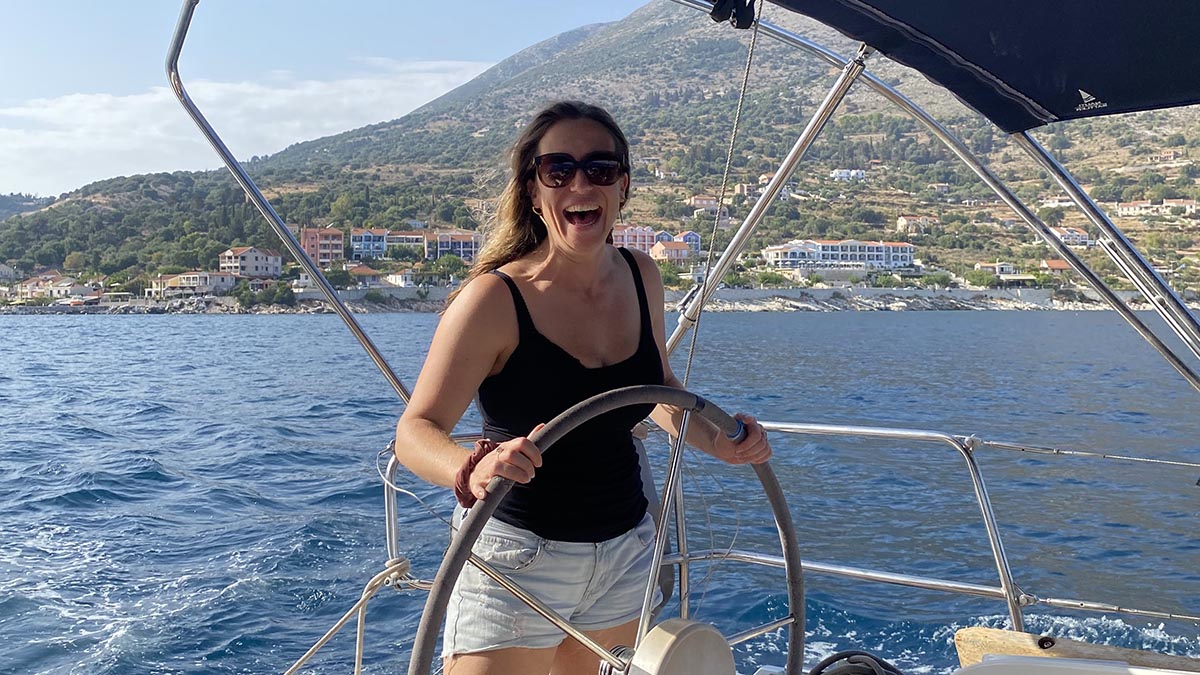 Happy woman steering a sailboat, smiling widely while enjoying sailing on clear blue waters with a picturesque coastal village and lush mountains in the background on a sunny day