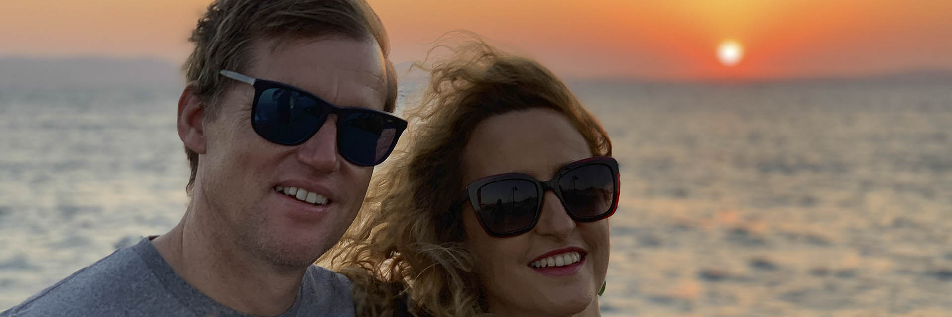 Close-up portrait of a smiling couple wearing sunglasses, enjoying a romantic sunset by the sea with warm golden hues in the sky, creating an intimate vacation moment