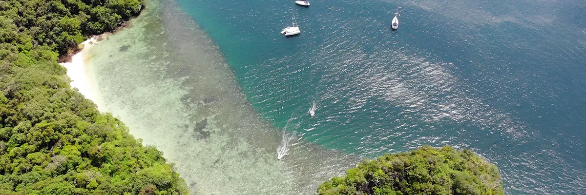 Aerial view of a secluded tropical bay with lush green vegetation, pristine clear waters, a sandy beach, and sailboats anchored nearby, creating a perfect sailing destination scene